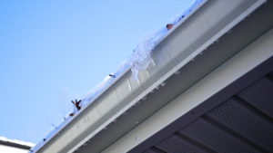 Close up of a white gutter on a house with snow, ice, and debris inside it. Ice is melting down the side of the gutter.